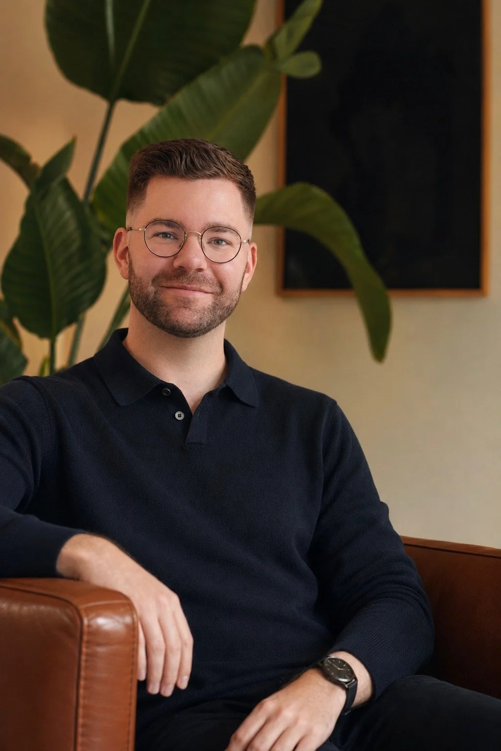 A man with glasses and a beard smiling, sitting on a leather chair with a large green plant and a blackboard in the background.