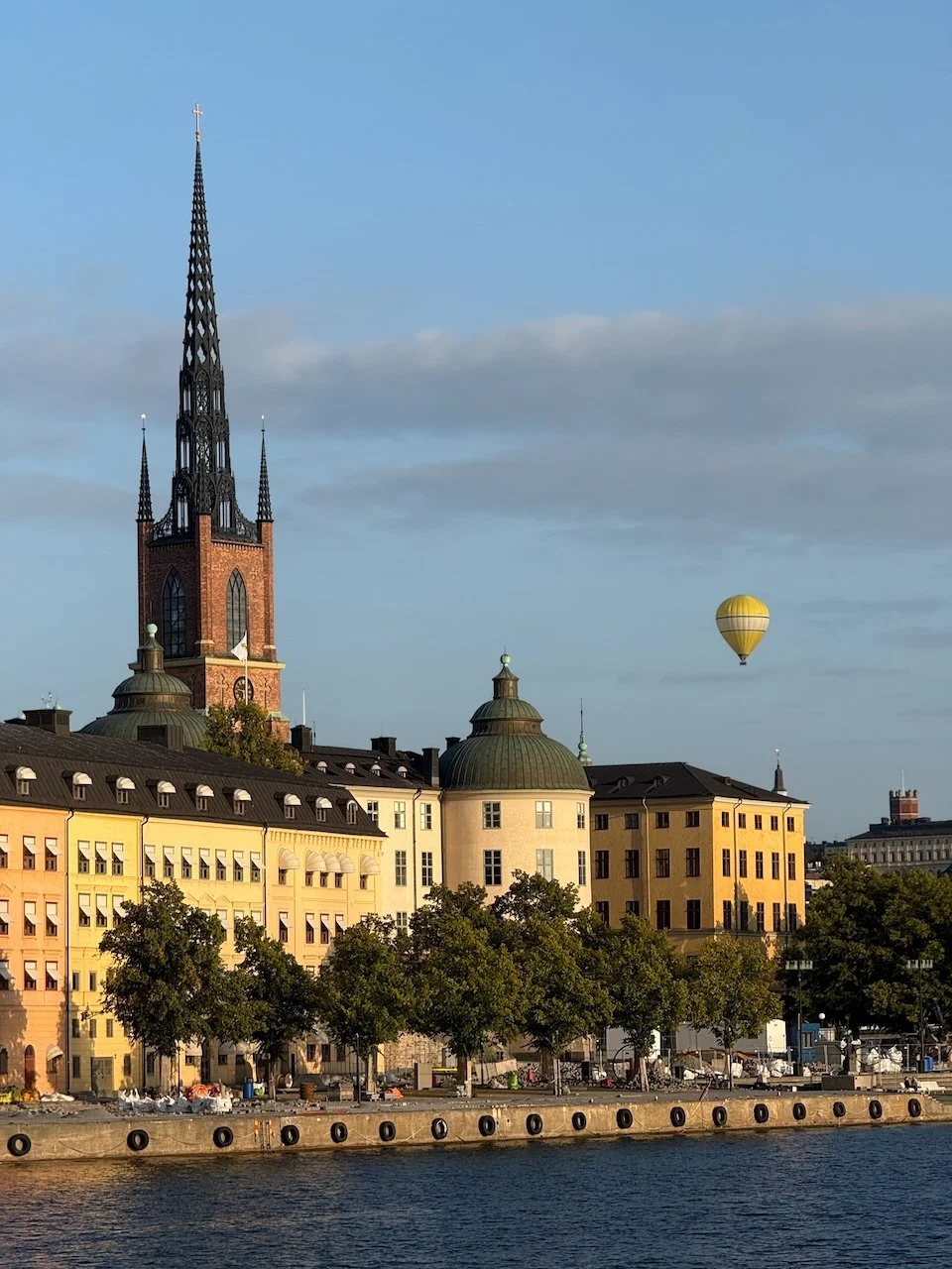 Cityscape with historic buildings along water, church with tall spire, and a yellow hot air balloon in the sky.