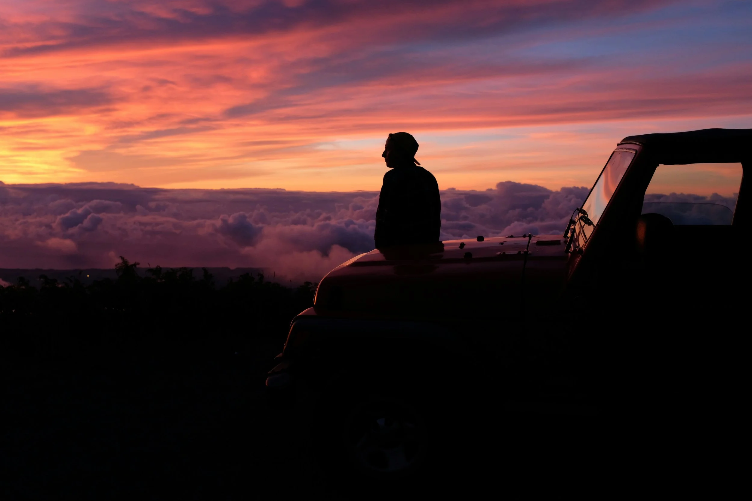 Silhouette of a person sitting on the hood of a car, overlooking a colorful sunset sky above clouds.