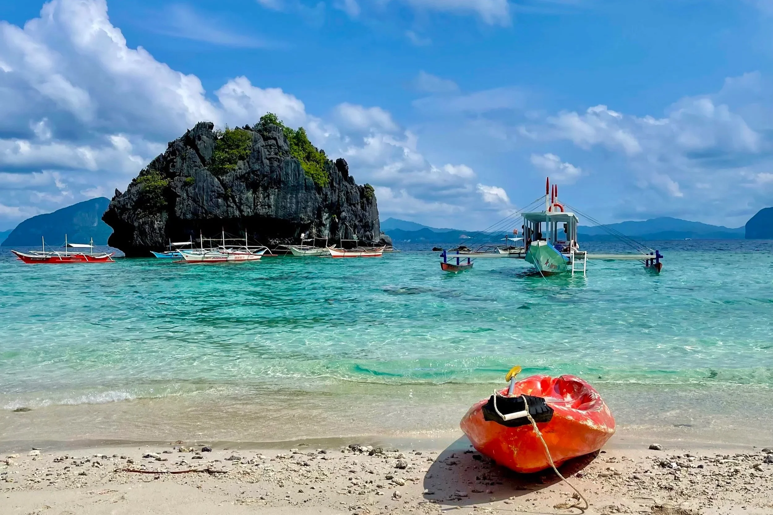 A tropical beach with clear turquoise water, a rocky island with greenery, several small boats anchored near the shore, and a red life raft in the sand with a yellow float, under a partly cloudy sky.