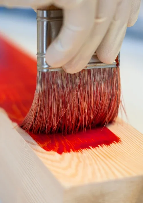 A paintbrush applying red colour to a wooden surface