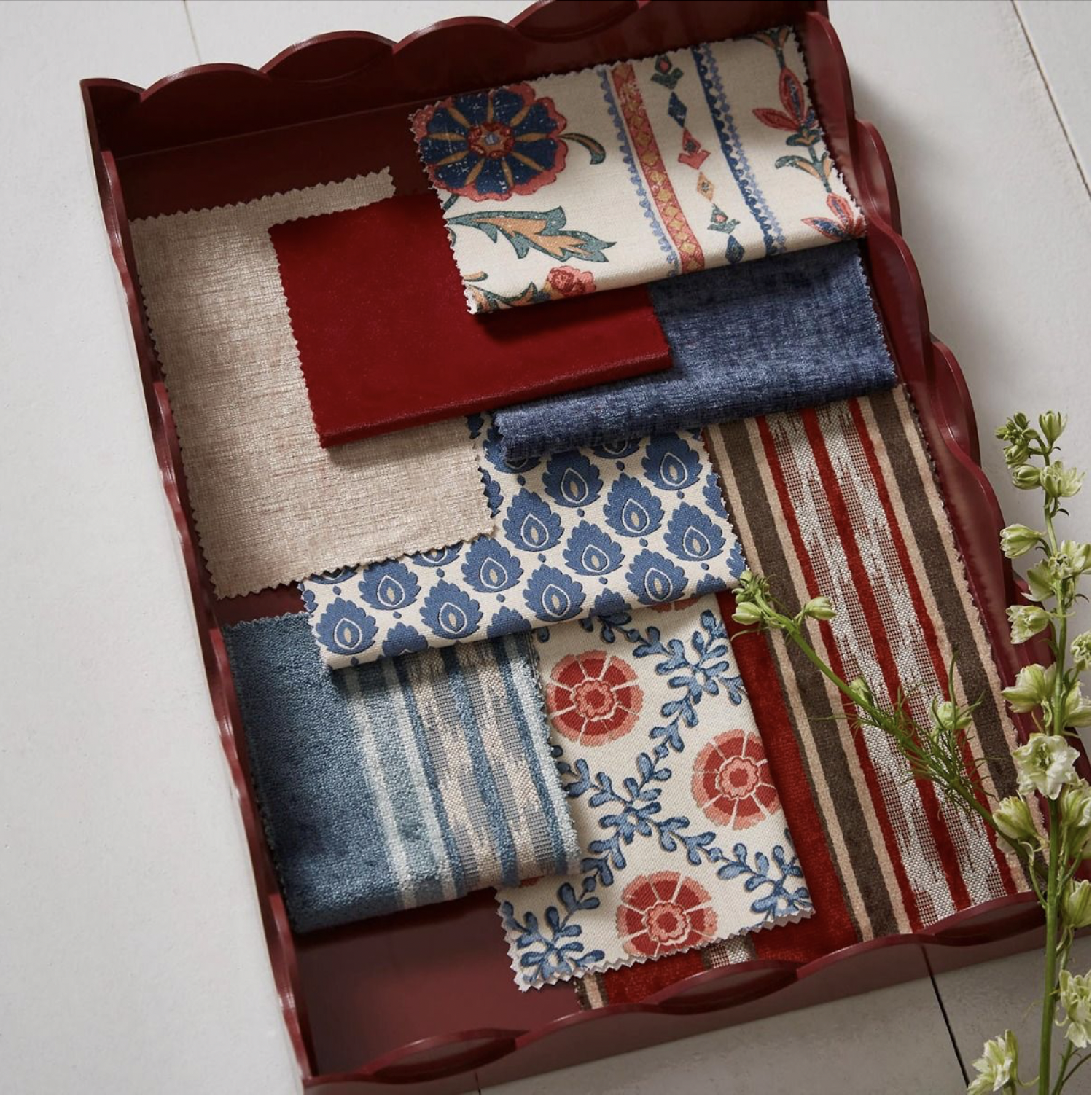 Stoffmuster in einer roten Schale auf weißem Hintergrund, verschiedene textile Stoffproben mit blau, rot, beige und floralem Design, teilweise gestrickt oder mit Mustern, daneben grüne Blumen.