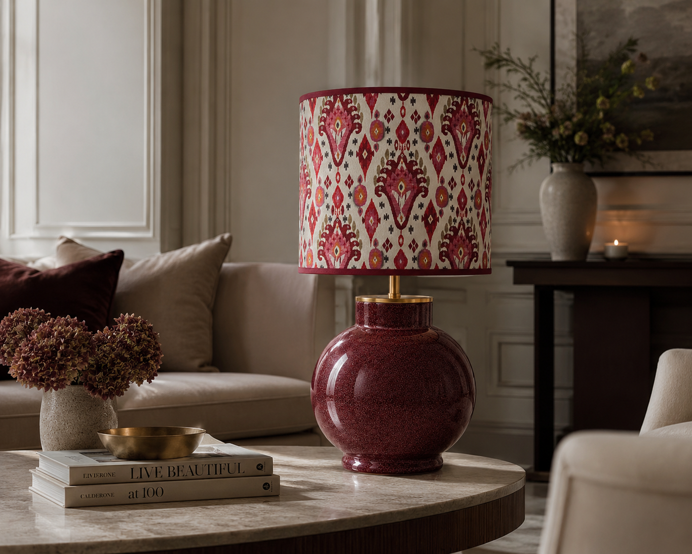 Ein runder Tisch mit einer Vase mit Hortensien, einem goldenen Teller und Büchern. Im Hintergrund eine Couch und eine Tischlampe mit einem roten, gemusterten Lampenschirm. Weiter hinten eine Vase mit Blumen auf einer Konsole und eine brennende Kerze.