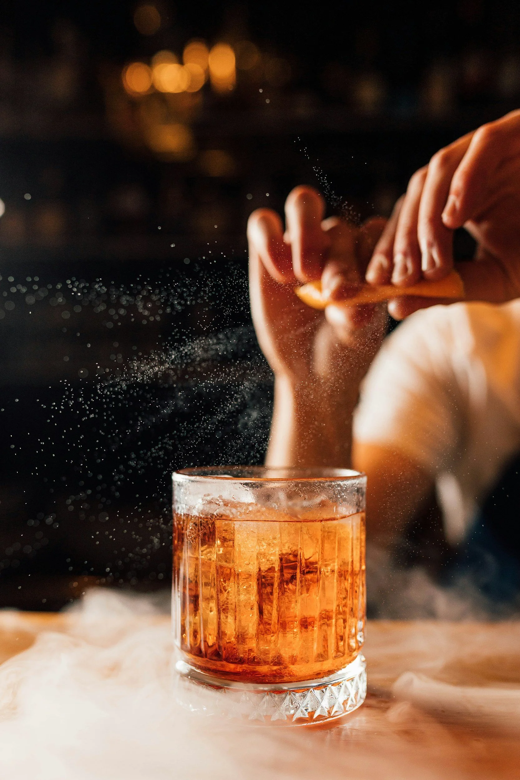 A person's hand is seen sprinkling salt into a glass of orange-colored beverage on a wooden surface, with a blurred dark background and warm bokeh lights.