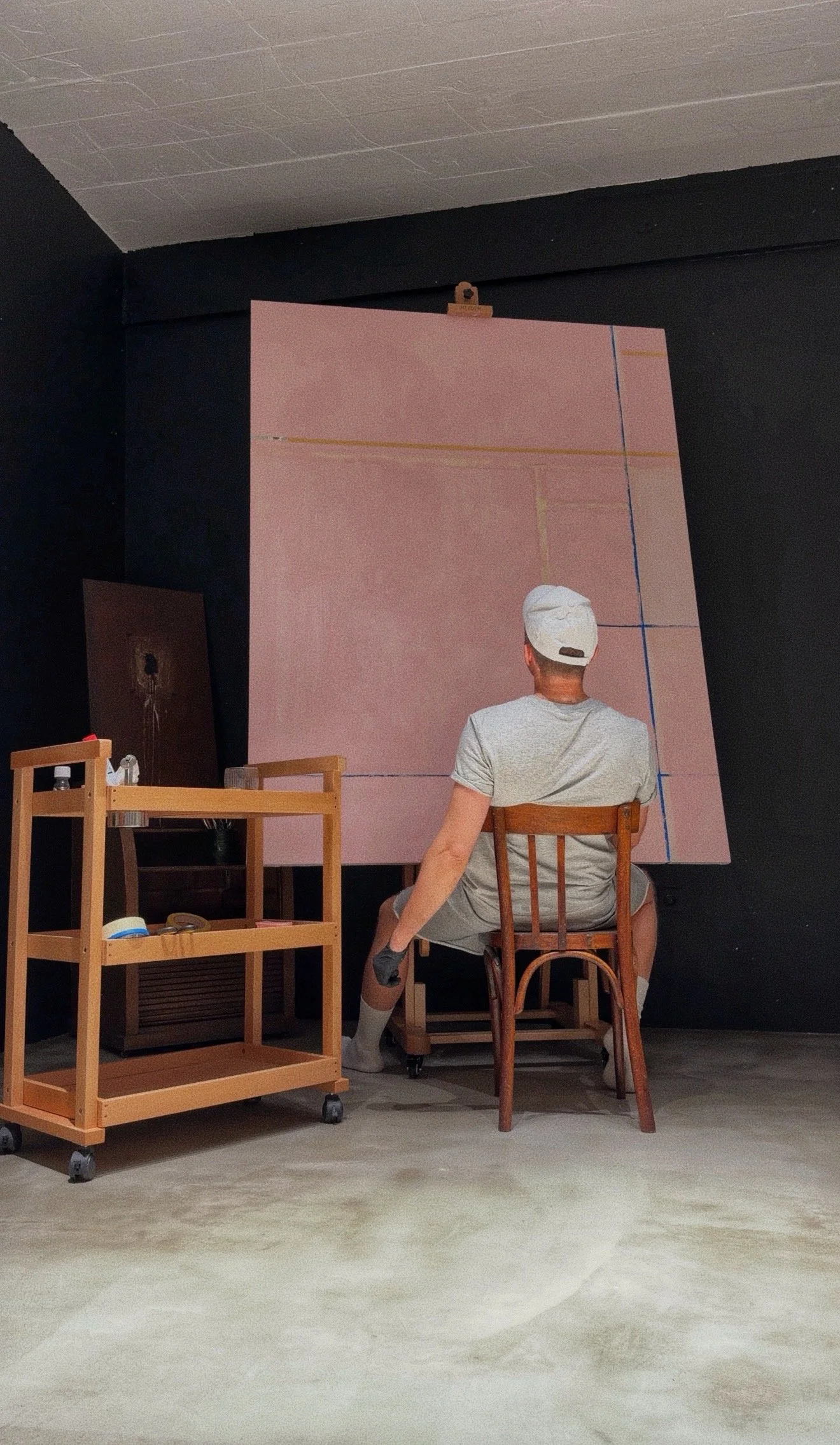 A man with a white cap and gray t-shirt sitting on a wooden chair, facing a large pink canvas with faint lines, in an art studio with black walls and a wooden shelving unit.