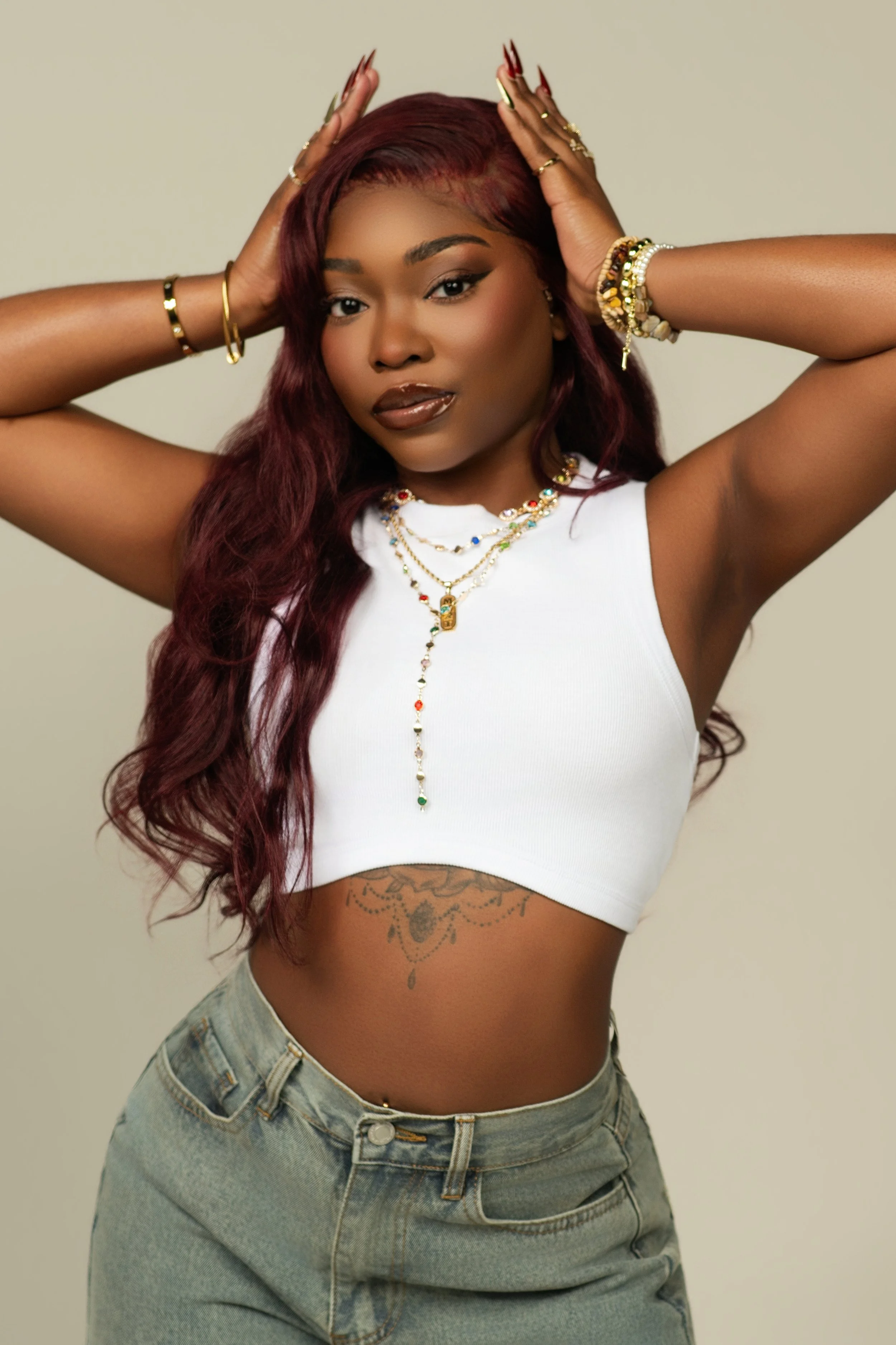 A woman with long wavy burgundy hair, wearing a white crop top, jewelry, and light blue jeans, posing with hands behind her head against a plain background.