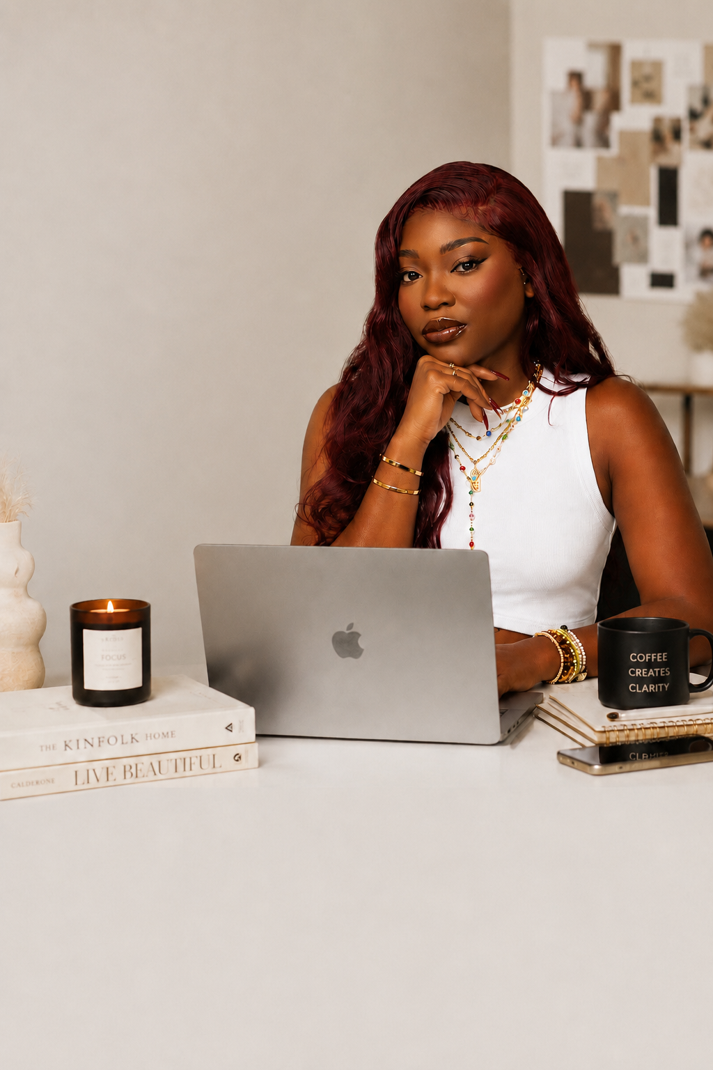 A woman with long red curly hair sitting at a white desk, working on a silver MacBook laptop. She is wearing a white sleeveless top and layered jewelry. On the desk are a black mug with the words "Coffee Creates Clarity," a closed notebook, a smartphone, and a lit candle. Behind her is a wall with photos and decor.