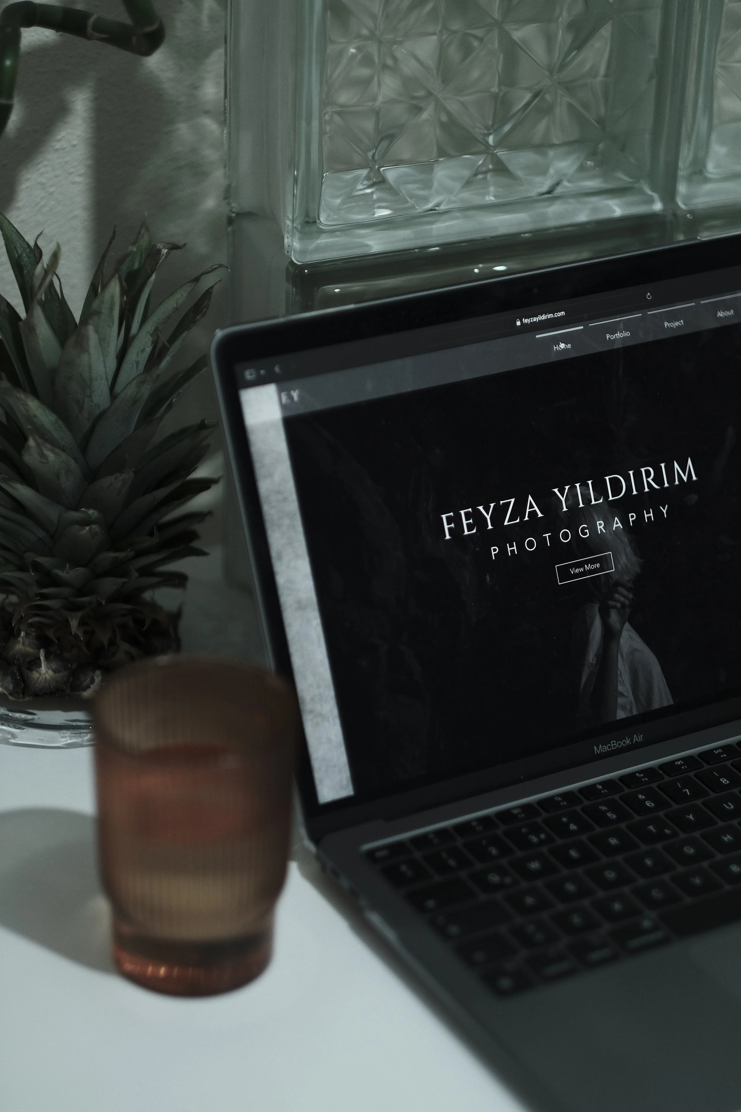 A close-up shot of a computer screen displaying a website titled 'Feyza Yıldırım Photography,' with a button labeled 'View More.' In the background, partly visible, there is a glass pineapple-shaped decorative item, a laptop keyboard, and a glass brick display. A person's finger is slightly blurred in the foreground.