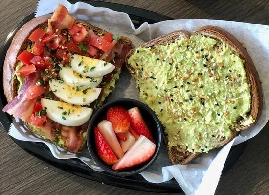 A breakfast tray with avocado toast topped with black sesame seeds, an open-faced bacon and tomato sandwich with chopped herbs, a side of sliced strawberries, and a small cup of apple slices with strawberry pieces.