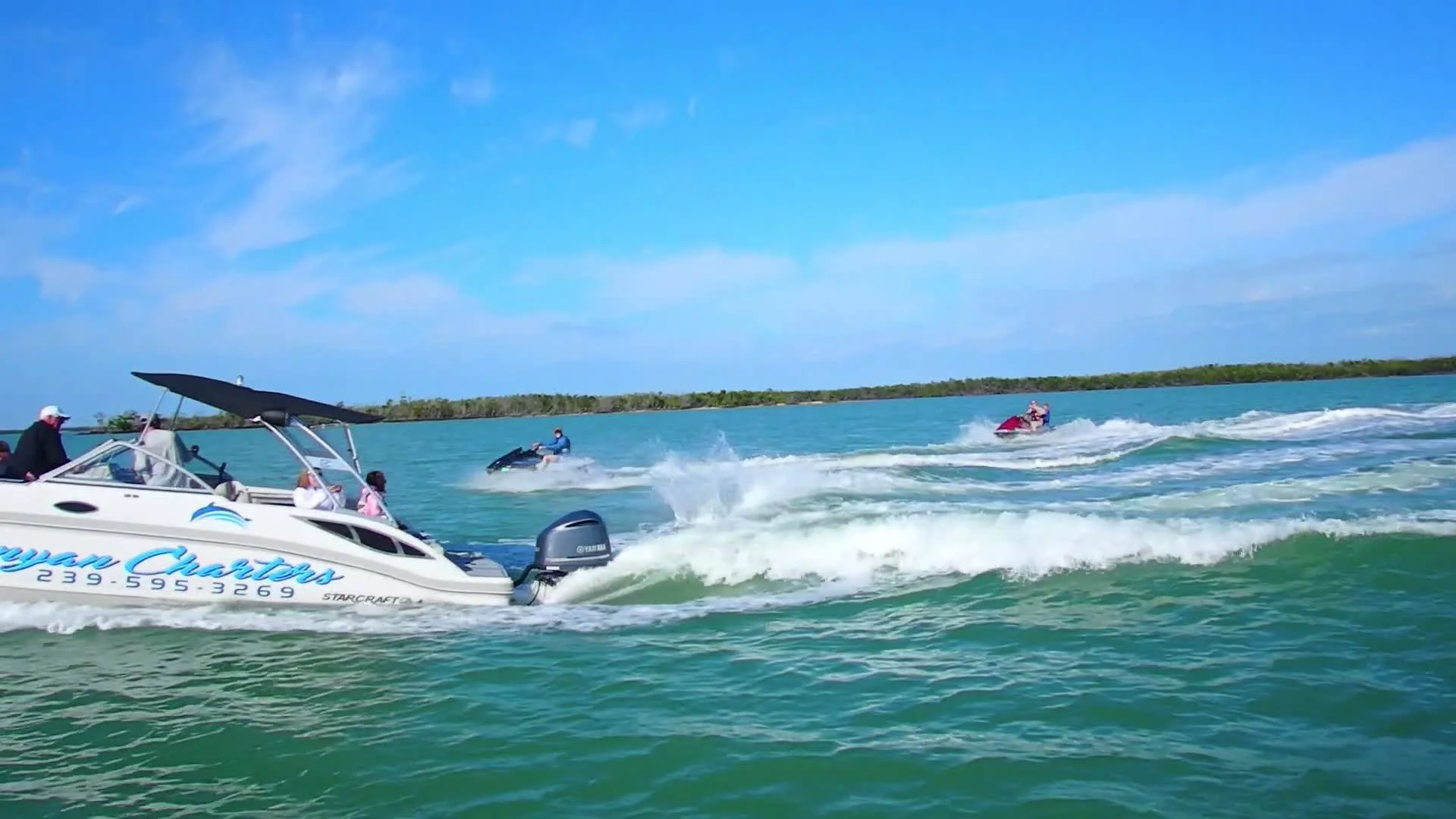 People enjoying water activities with a white boat and jet skis on blue water, with a distant land and sky with wispy clouds.