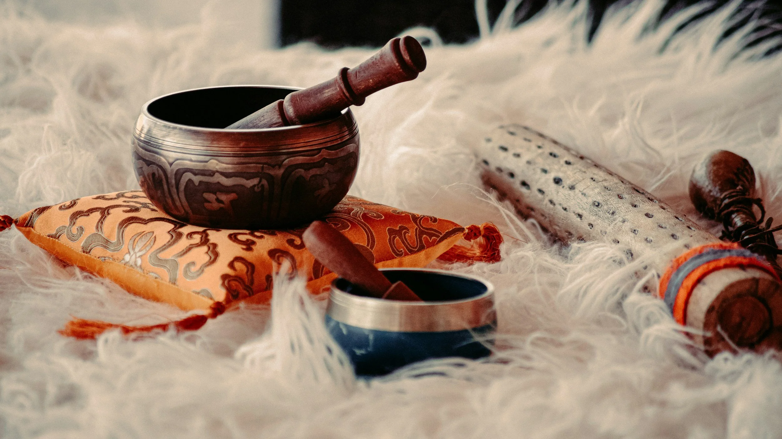 A Tibetan singing bowl with a wooden striker inside, placed on an ornate orange and gold cloth, surrounded by a Himalayan salt lamp, a small black dish, and colorful prayer beads, all on a fluffy white surface.