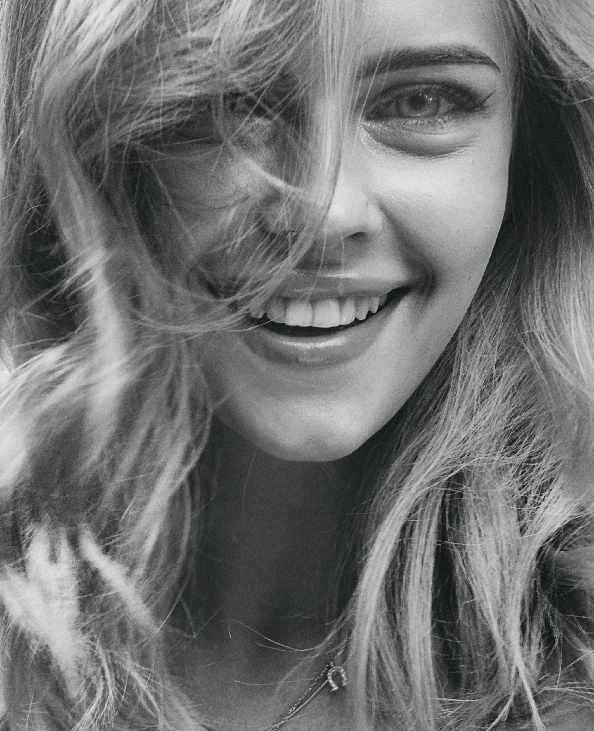 Black and white close-up photo of a smiling woman with long, wavy hair covering part of her face, featuring a sparkle piercing on her nose and a chain necklace with a key pendant.