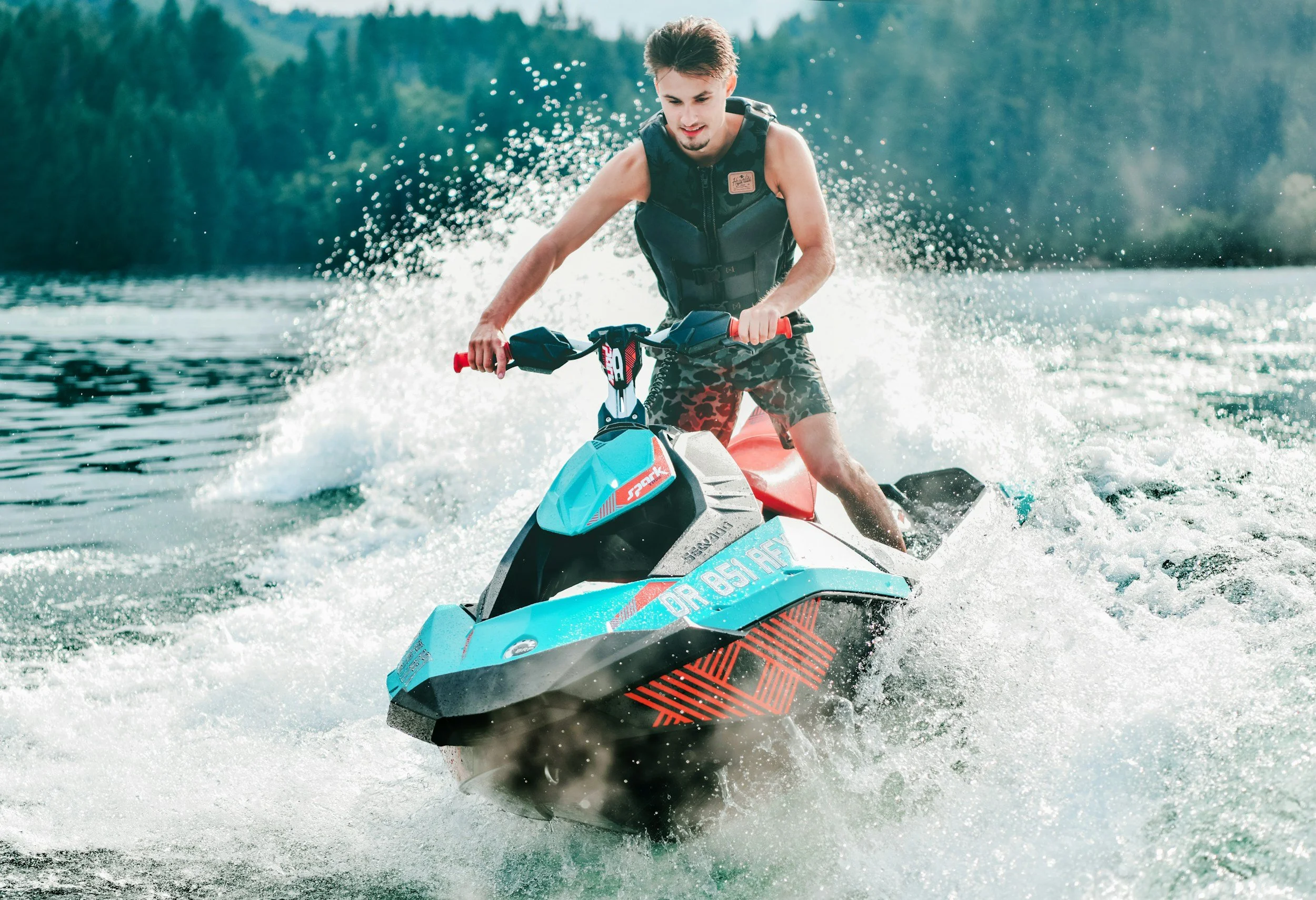 A young man riding a jet ski on a lake surrounded by trees. He is wearing a life jacket and camouflage shorts, and he appears to be enjoying the water activity.