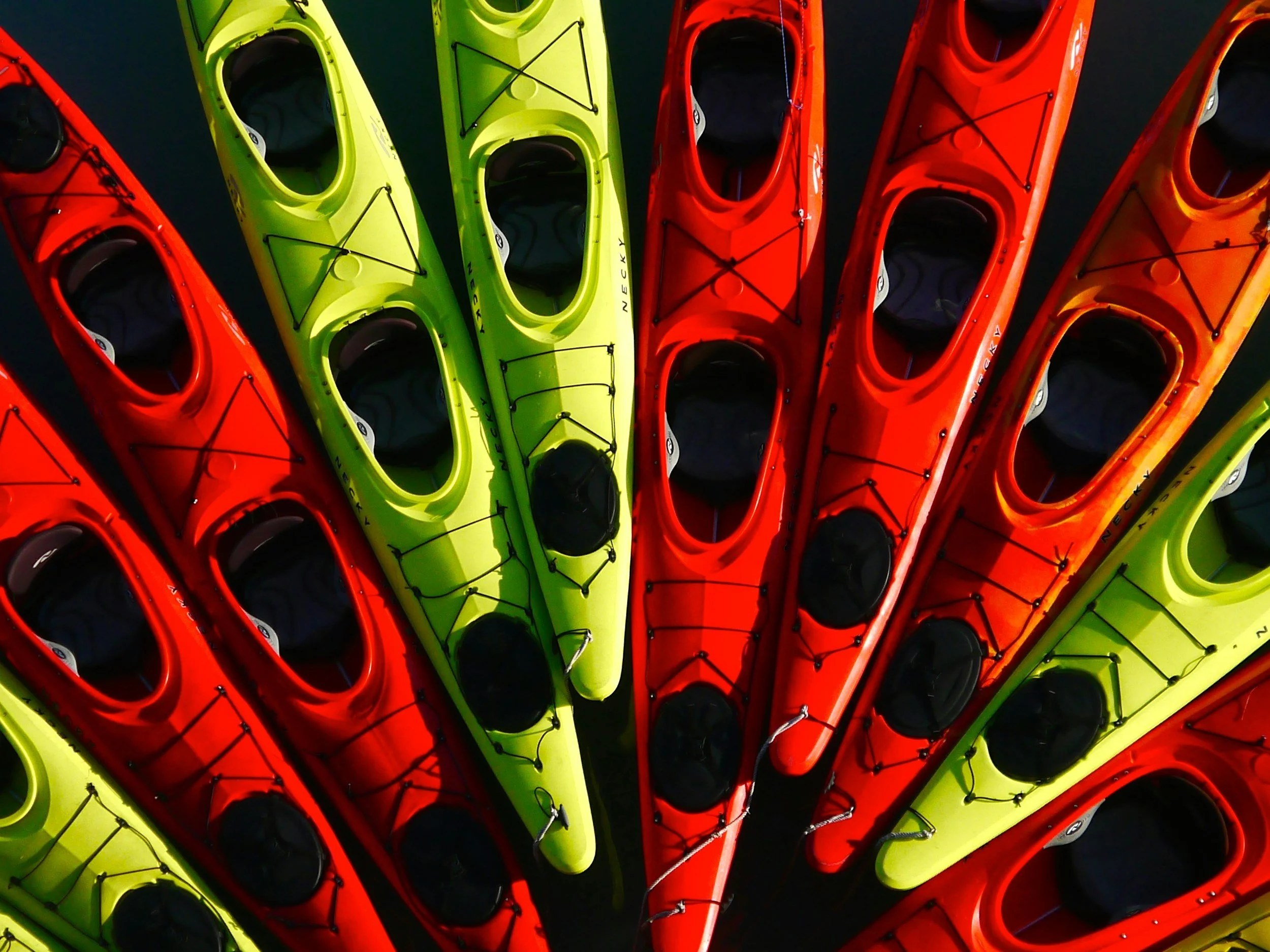 A colorful collection of kayaks arranged in a pattern, including red, yellow, and orange kayaks with black interior and rigging.