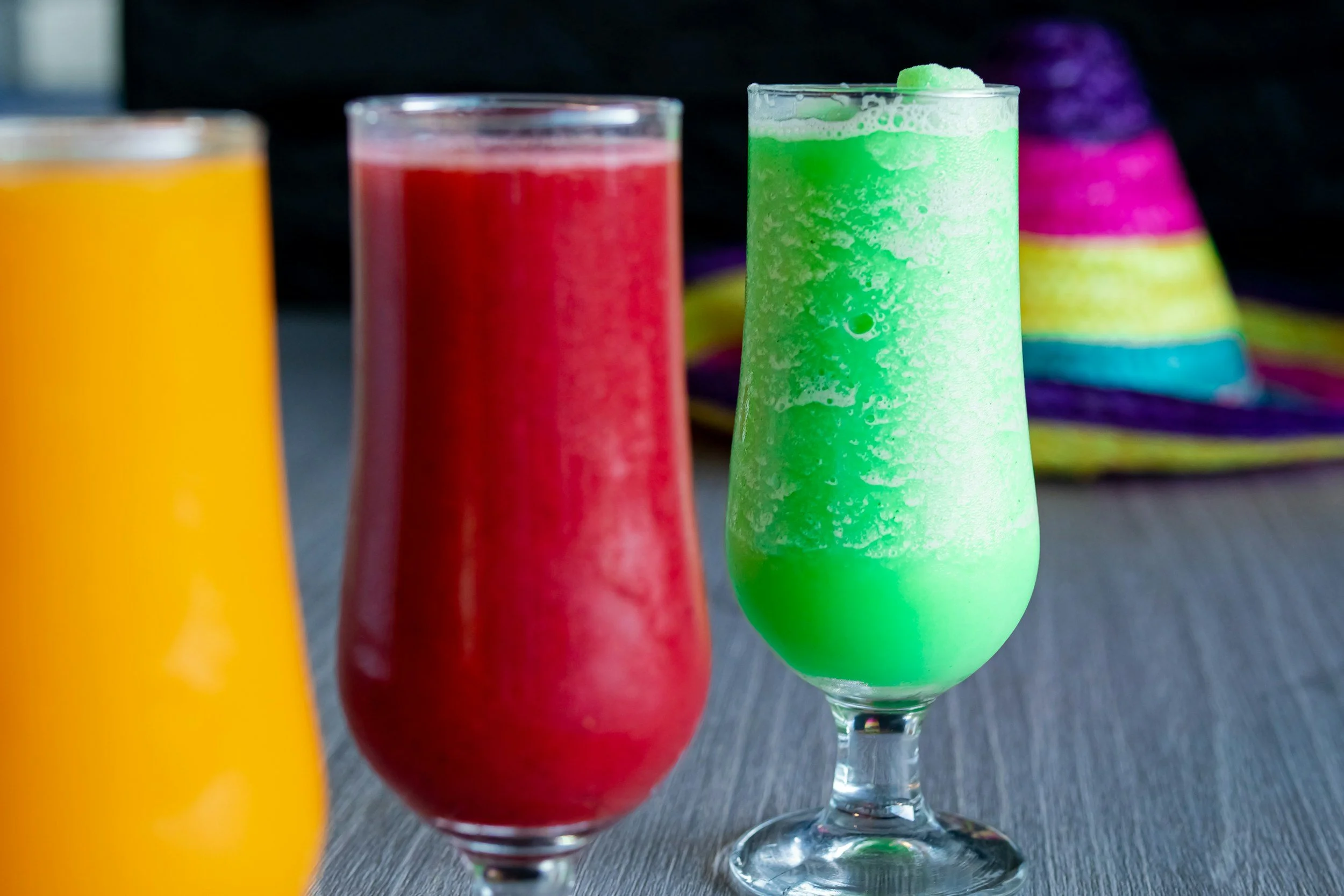 Three colorful smoothies in tall glasses on a dark surface, with a blurred rainbow-colored cloth in the background.