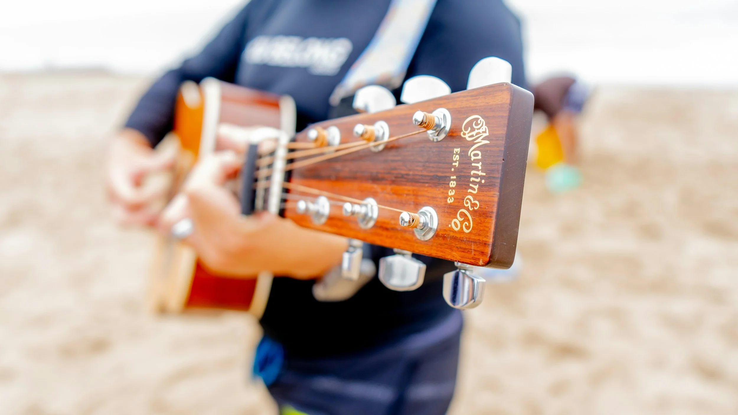Close-up of a person holding a Martin acoustic guitar outdoors, with a sandy background and blurred figures in the distance.