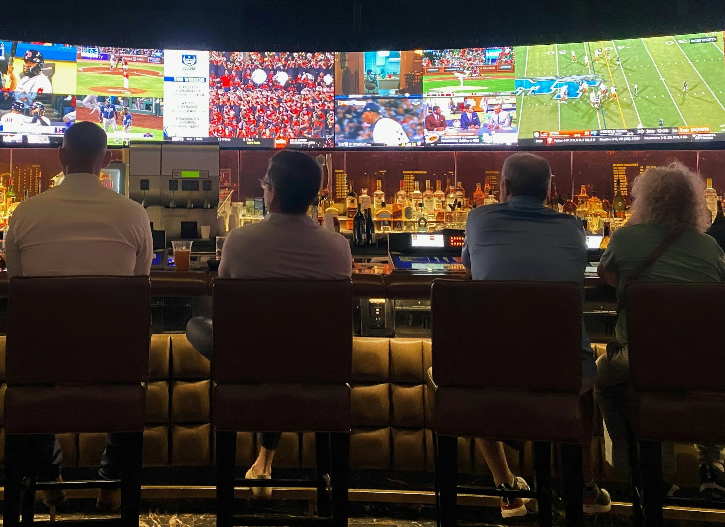 A sports bar with four people sitting at a counter, watching multiple large screens showing various sports events, including baseball and football. The bar area has bottles of alcohol and a beer tower.