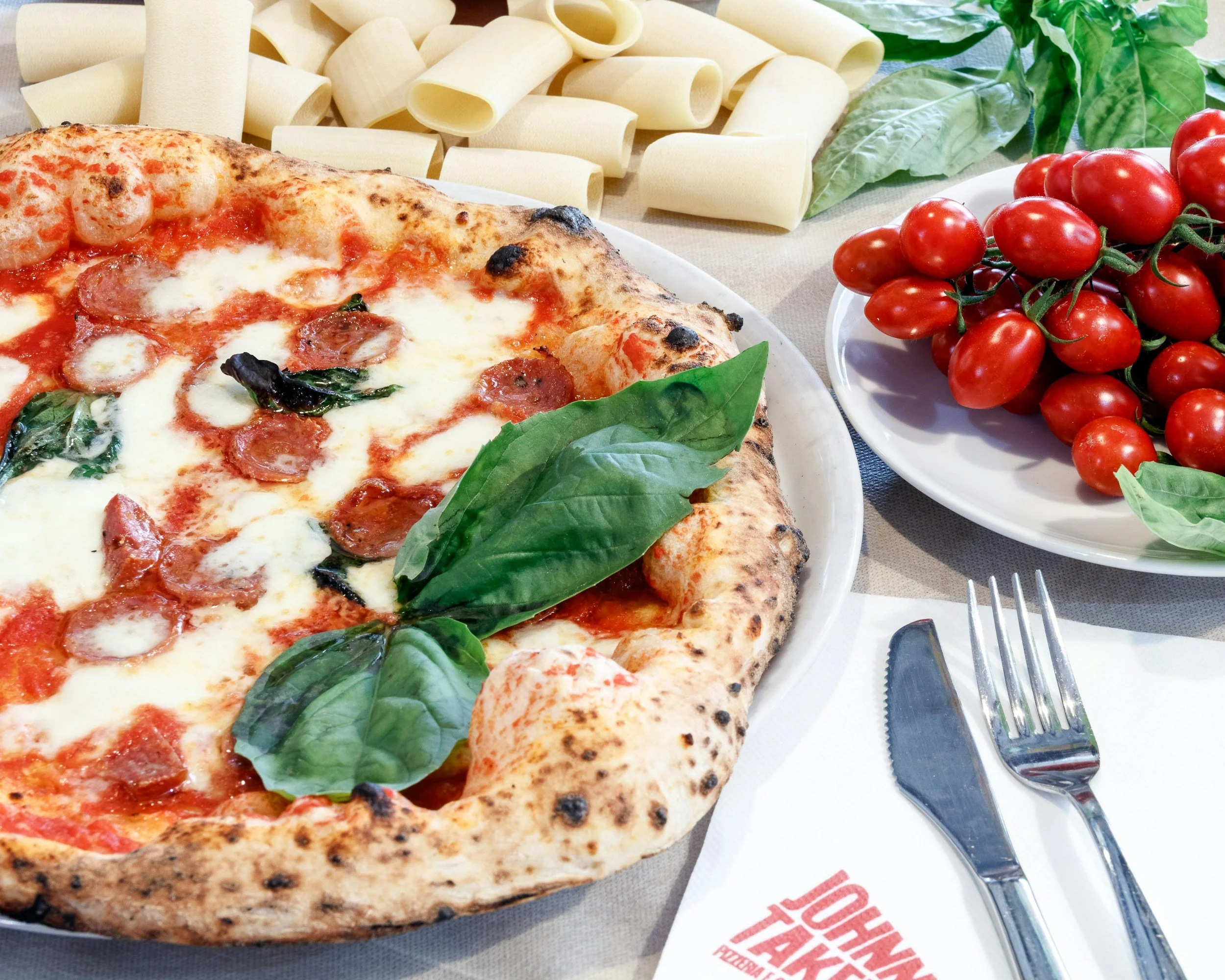 Close-up of a pepperoni pizza topped with basil leaves on a white plate with a knife and fork beside it, next to a bowl of cherry tomatoes and uncooked pasta and fresh basil leaves on a table.