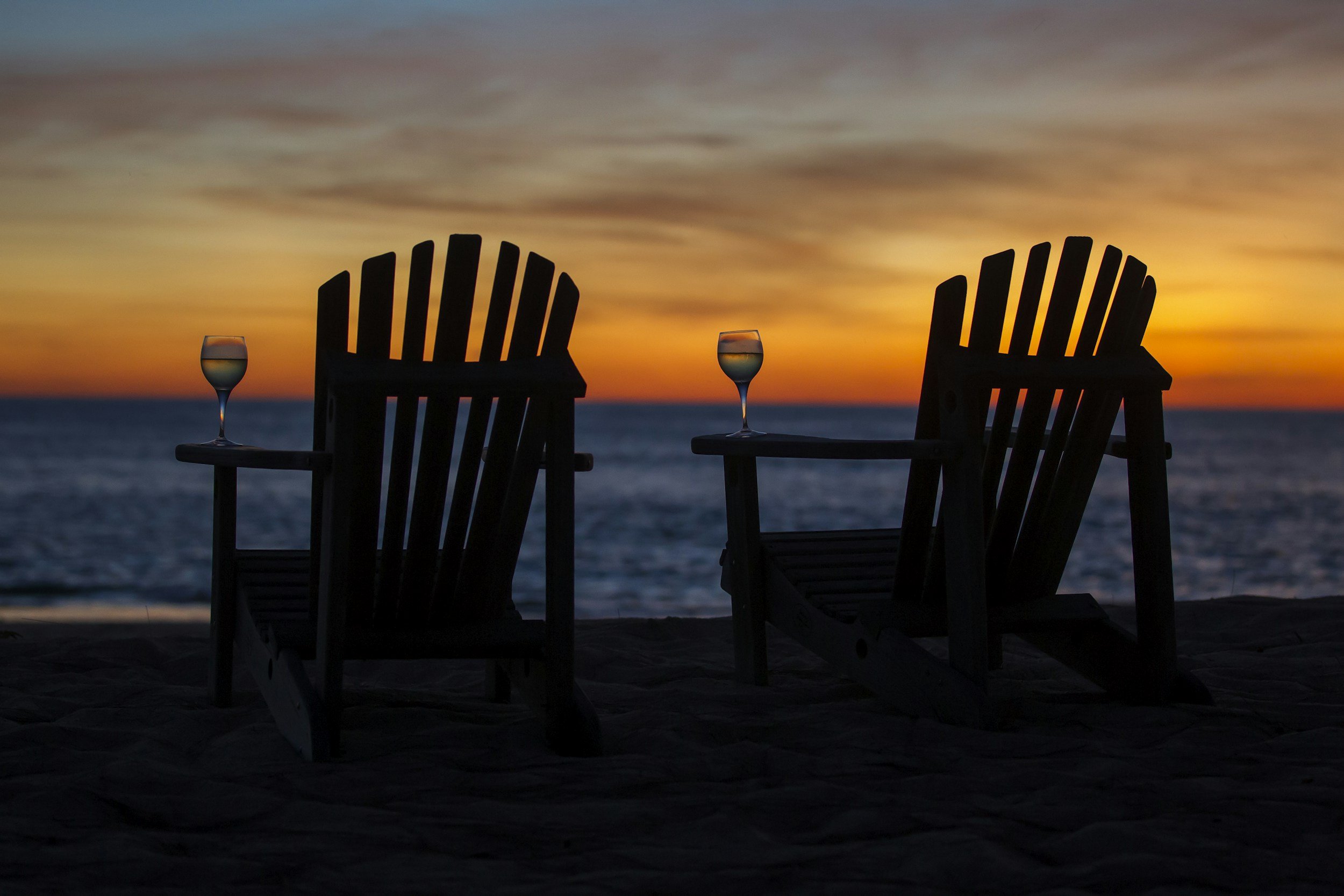 Silhouette of two Adirondack chairs on the beach at sunset with two wine glasses on the armrest, ocean in the background, and colorful sky with orange, yellow, and blue hues.