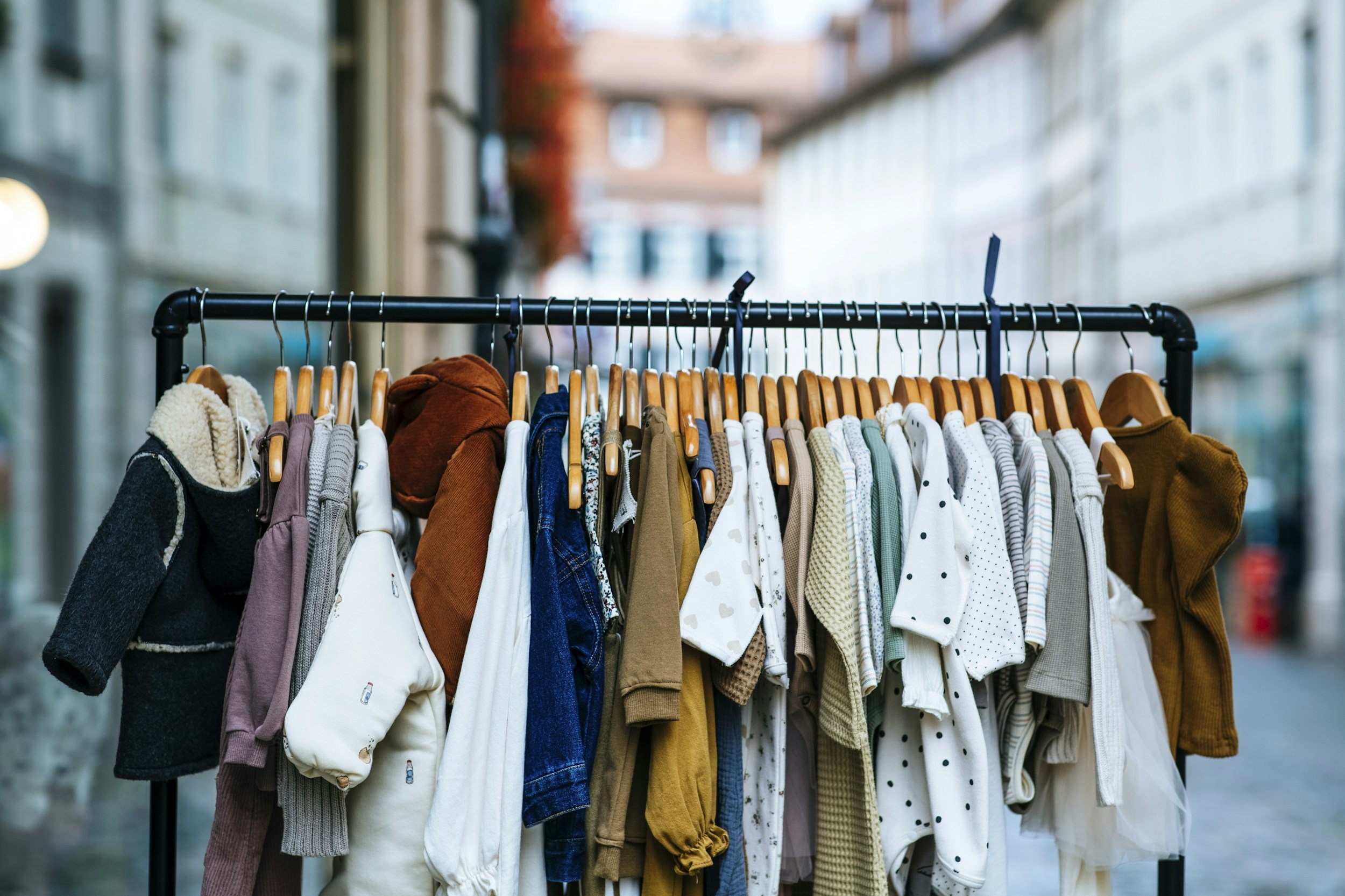 A clothing rack outdoors with various children's jackets and sweaters hanging on wooden hangers, with a city street background.