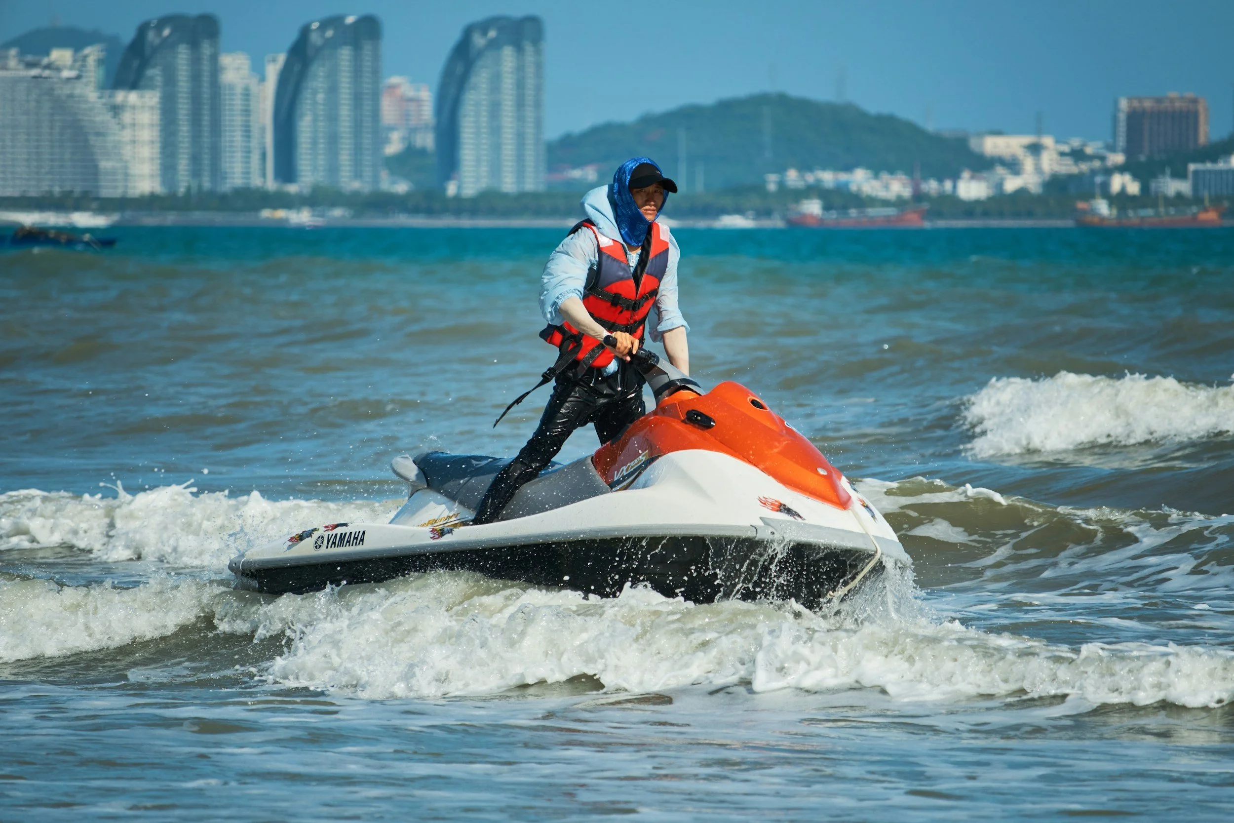 Person wearing a life jacket riding a Yamaha jet ski on the water near a cityscape with tall buildings and a hill in the background.
