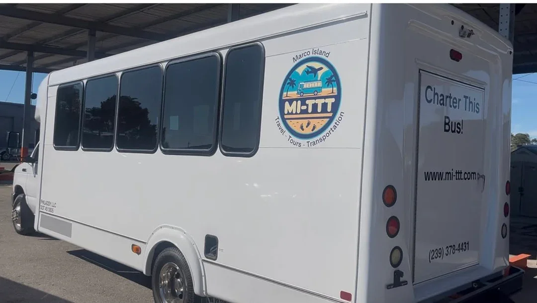 White bus with a logo on the side that says 'Marco Island MI-TTT' with a graphic of a bus and palm trees, advertising charter services for travel, tours, and transportation.