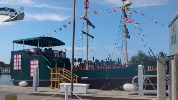A pirate ship-themed boat decorated with colorful flags, docked at a marina with a clear sky in the background.