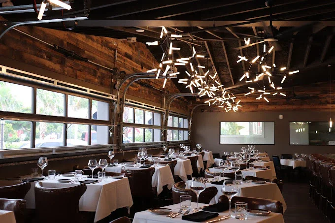 Upscale restaurant dining area with white tablecloths, wine glasses, and cutlery, featuring a modern light fixture and large windows showing an outdoor view.