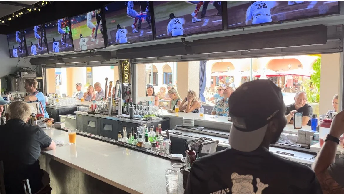 Inside a sports bar with a bar counter in the foreground, patrons sitting at the bar and outside, watching sports on multiple large screens showing a baseball game. The interior has bottles and glasses on the bar, and outdoor seating is visible through large windows.
