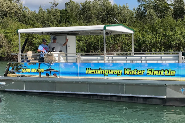 A pontoon boat labeled 'Aemingway Water Shuttle' with two people on board, one waving, on a body of water with trees in the background.