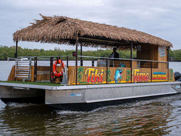 A floating tiki hut restaurant on the water with a thatched roof and colorful signage that reads 'Tiki Time' and features a cartoon character wearing a Hawaiian shirt and sunglasses.