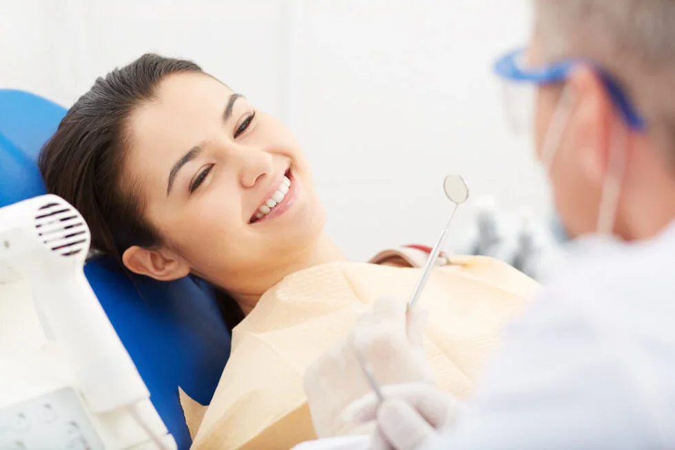 A woman with a big smile lying in a dental chair during a checkup, with a dentist holding dental tools near her mouth.