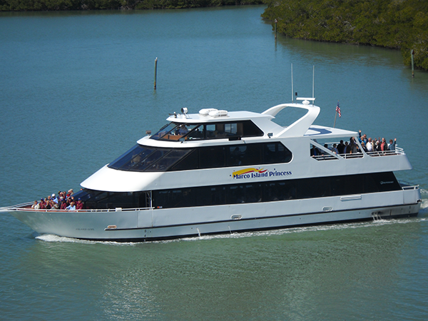 A large yacht named Marco Island Princess sailing on a river with green trees in the background, carrying passengers.