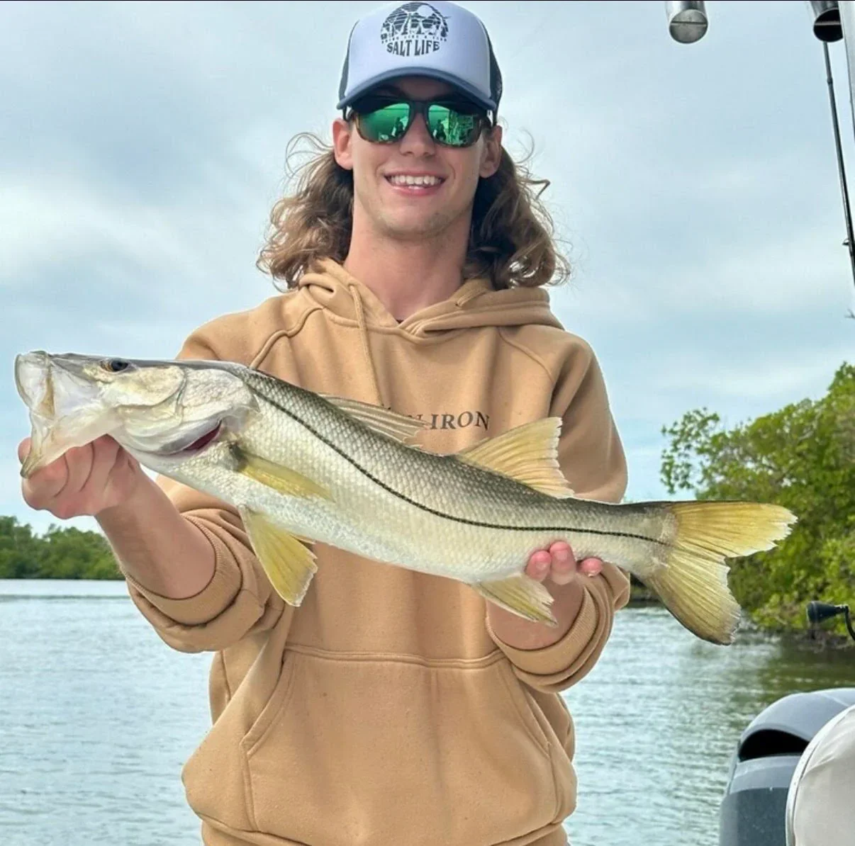Man smiling and holding a large fish near a body of water, wearing a beige hoodie, a cap with 'Salt Life' logo, and sunglasses. Trees and a cloudy sky in the background.