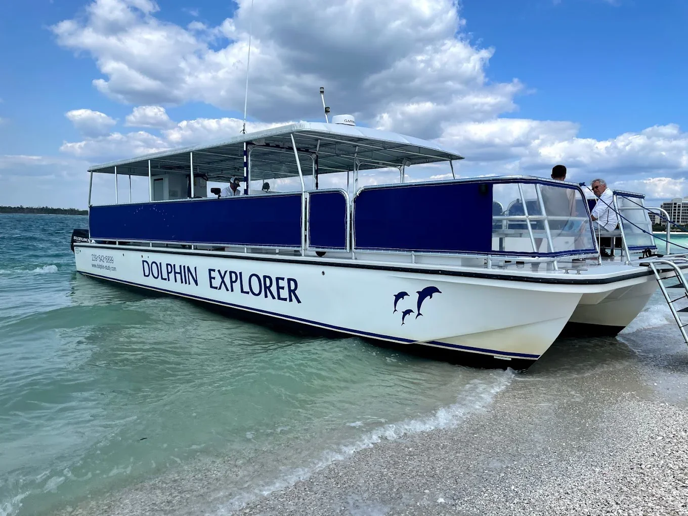 A white and blue dolphin-shaped boat named Dolphin Explorer on the water near a sandy beach, with a few people on board and a cloudy sky overhead.