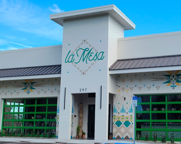 Front of a building named La Mesa with colorful geometric and native-themed decorations, green window frames, and a sidewalk with a handicapped parking sign.