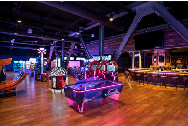 An arcade with various gaming machines, including a colorful air hockey table, a racing game, and other electronic arcade games in a dark, modern space with wooden floors and ambient lighting.