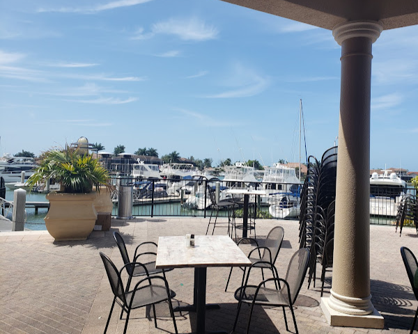 Outdoor patio with chairs around a table, overlooking a marina filled with yachts, under a partly cloudy sky.