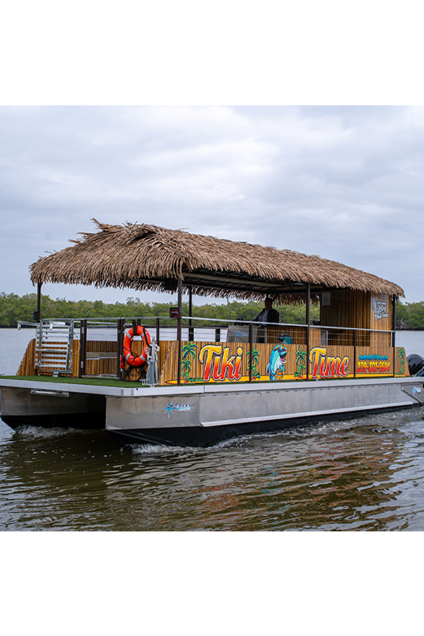 A houseboat with a thatched roof cruising on a body of water, with the words "Tiki Time" and tiki-themed decorations on its side.