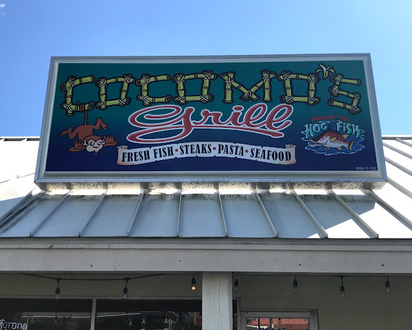 Signboard for a restaurant named "Coconut Grove Grill" offering fresh fish, steaks, pasta, and seafood, with a cartoon coconut and a crab mascot.