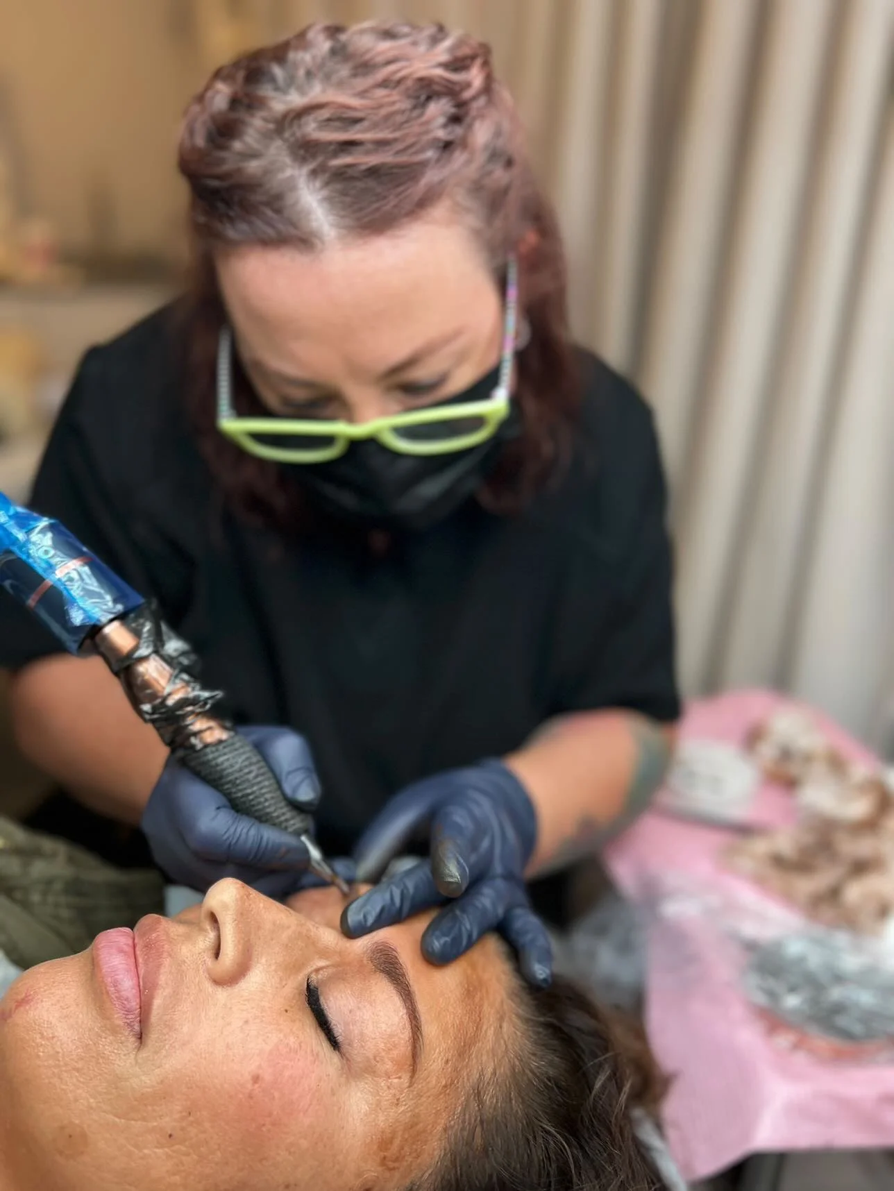A woman with glasses and a black face mask receives a tattoo on her face from a tattoo artist wearing black gloves and a black shirt.
