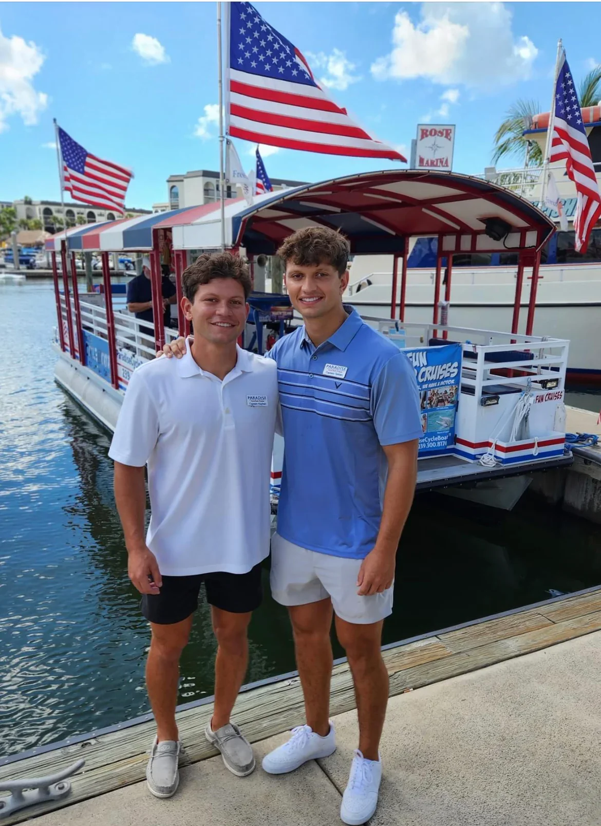 Two young men standing on a dock in front of a paddle boat decorated with red, white, and blue stripes and American flags. The man on the left wears a white polo shirt and black shorts, the man on the right wears a blue polo shirt and white shorts. The background features a marina with boats and buildings, under a partly cloudy sky.