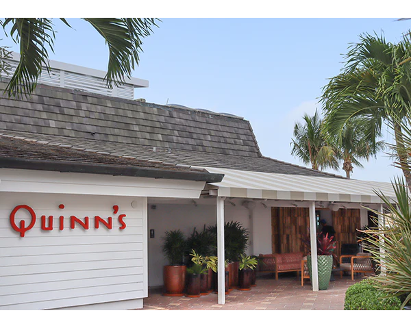 Exterior view of Quinn's restaurant with outdoor seating, potted plants, palm trees, and a covered patio area under a cloudy sky.