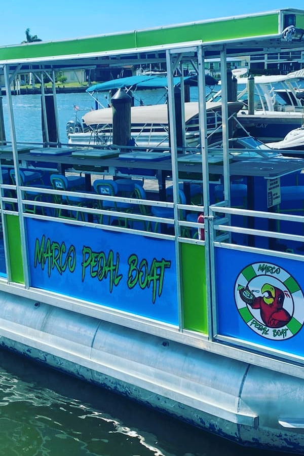 A pontoon boat named 'Marco Pedal Boat' floating on water, with life jackets stored on it, and multiple boats visible in the background docked at a marina.
