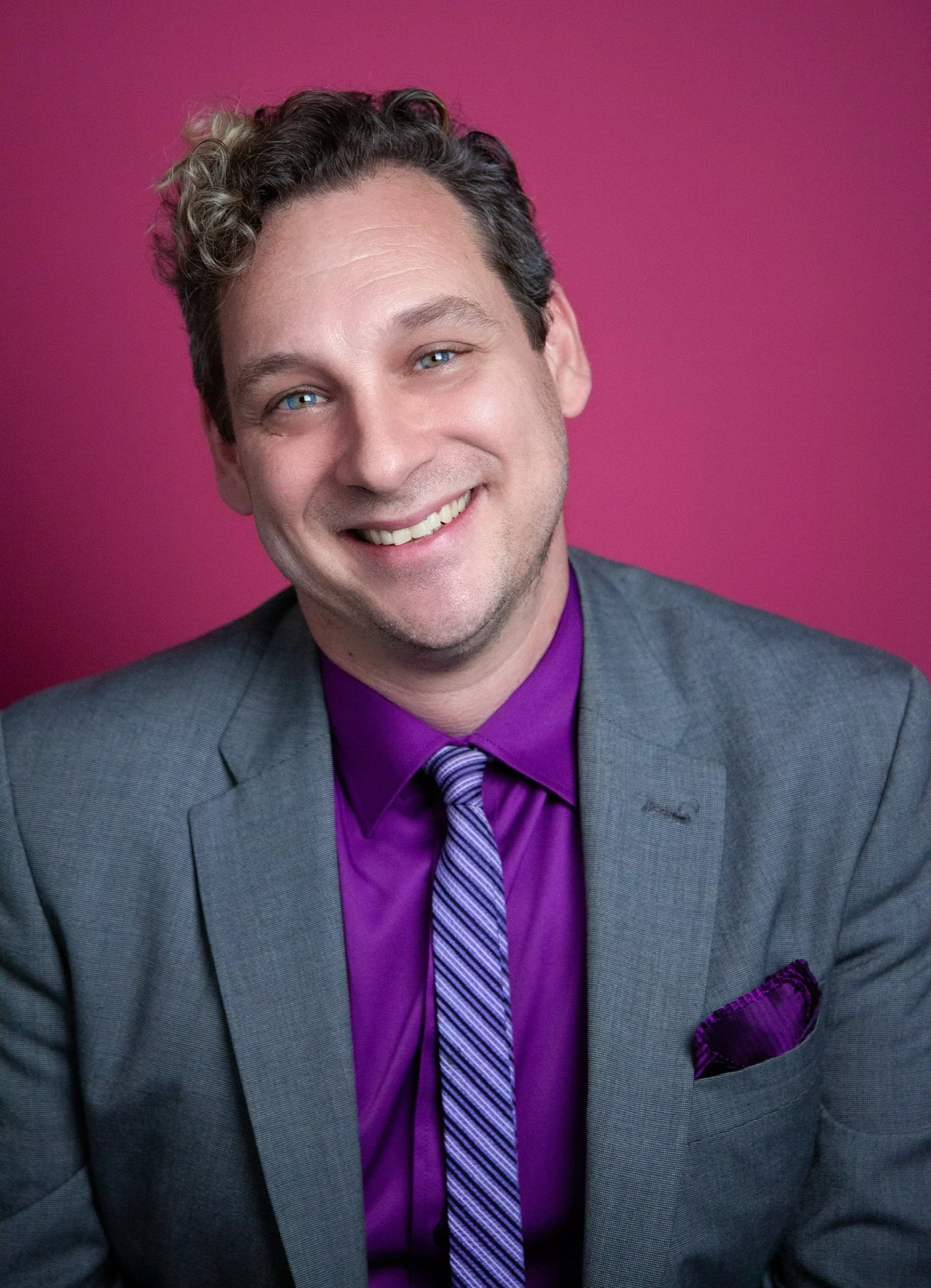 A man wearing a gray suit, purple shirt, and a striped purple tie, smiling against a pink background with a purple pocket square in his suit pocket.
