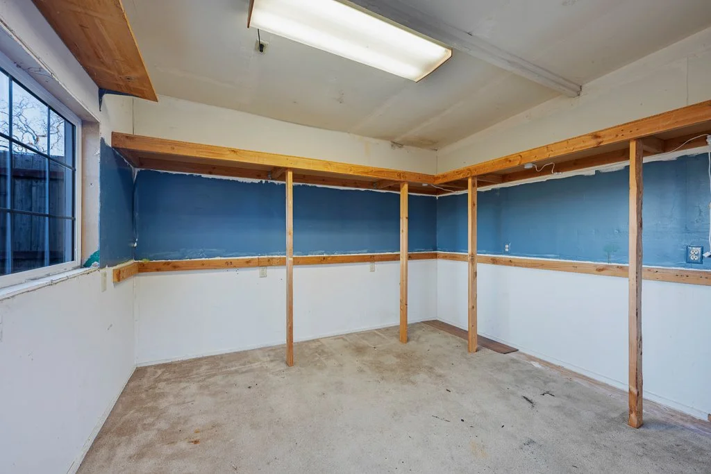 Empty room with exposed wooden beams, blue and white painted walls, a window on the left, and a beige carpeted floor.
