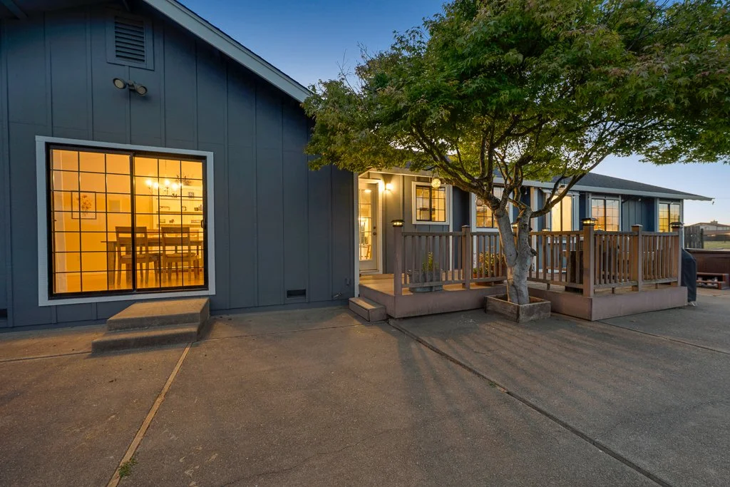 Exterior view of a house with a large window and a deck with a tree in front, lit during dusk.