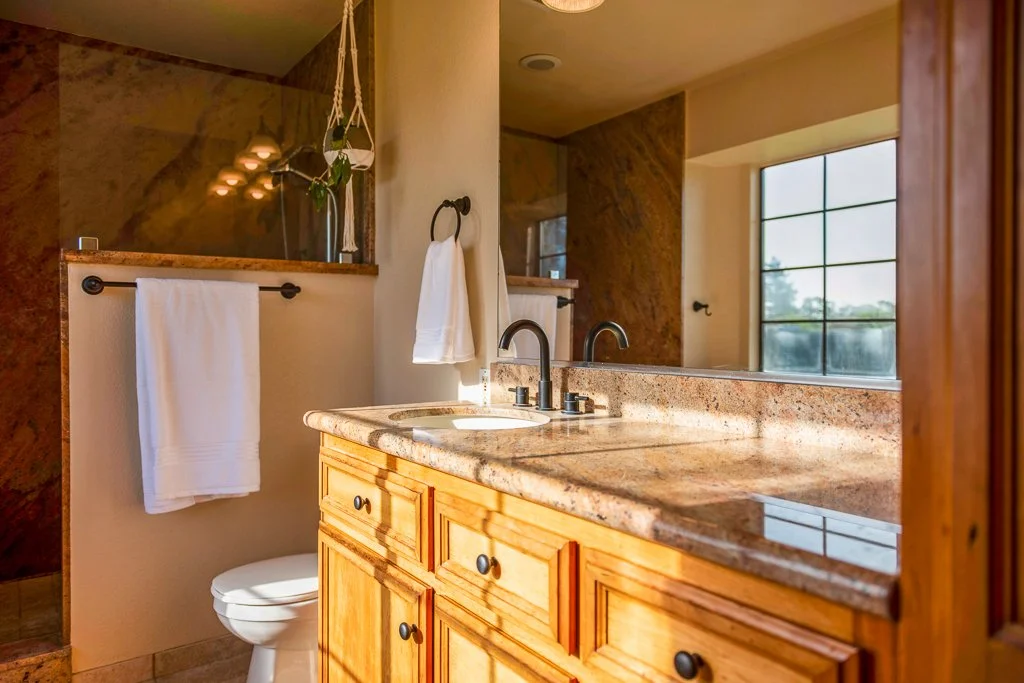 Bathroom with a wooden vanity, granite countertop, and double sink faucets. Towel rack with a white towel, a small white towel hanging from a ring, and a large window with grid patterns. A beveled glass mirror reflects a hanging plant and light fixtu