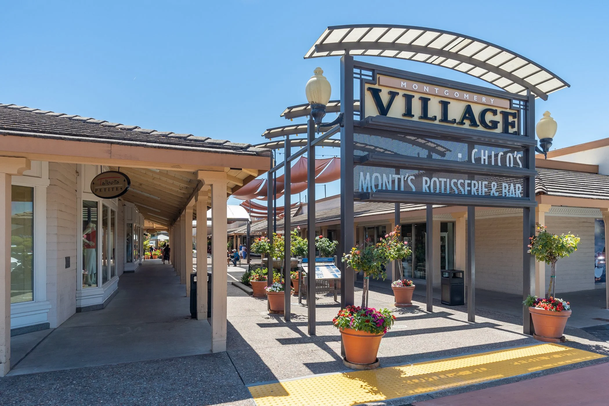 Outdoor shopping mall with storefronts and a large sign reading 'Montgomery Village' and 'Chico's, Monty's Rotisserie & Bar.' Potted plants and flowers decorate the area, under a clear blue sky.