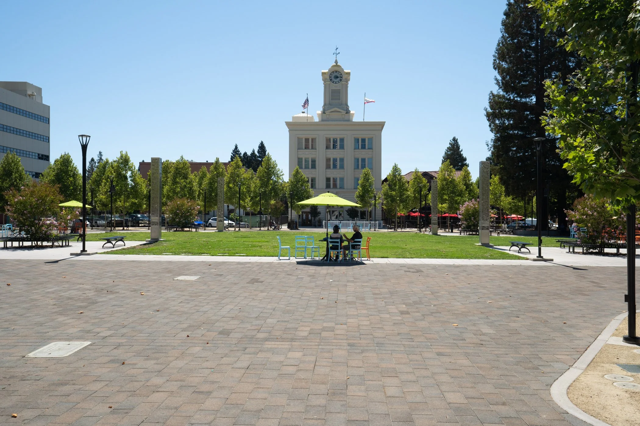 A city park with a large historic white building with a clock tower in the background. The park has a green lawn, trees, benches, and a few people sitting at a table with colorful chairs. The sky is clear and blue.